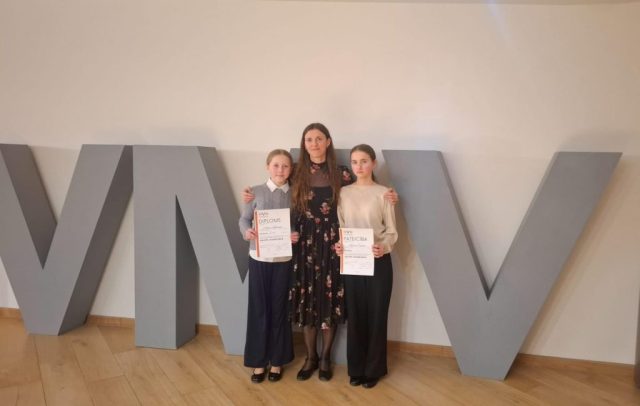 skolotāja Sunepa ar divām audzēknēm stāv pie Ventspils mūzikas vidusskolas zīmes iniciāļiem