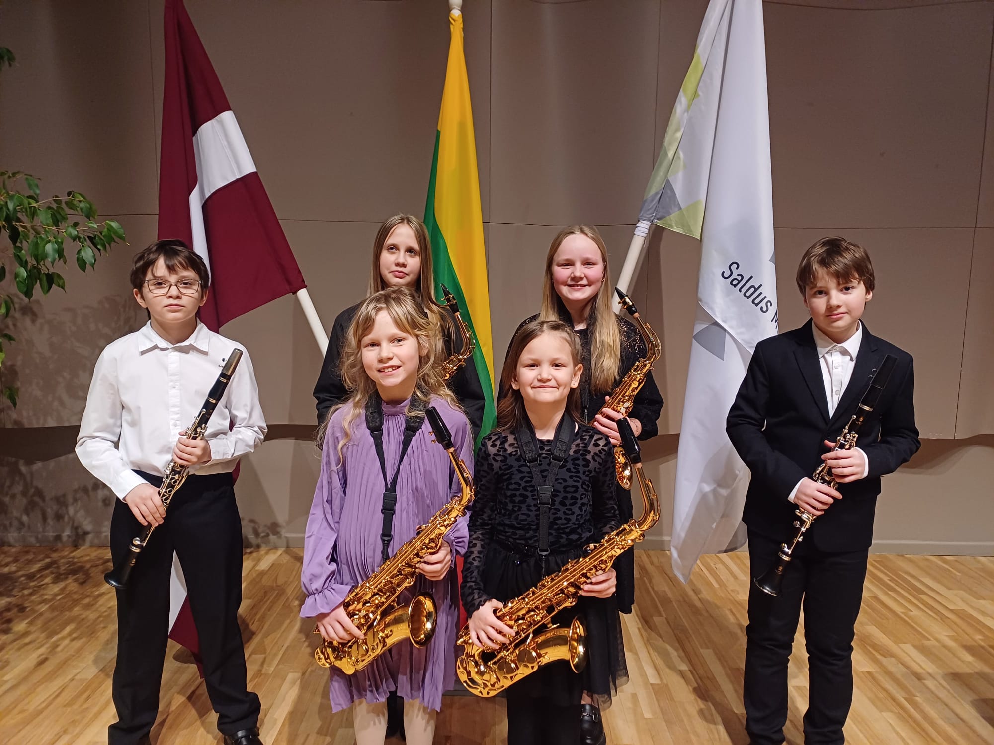 6 audzēkņi ar saksofoniem un klarnetēm. Pie Latvijas, Lietuvas un Saldus Mūzikas skolas karogiem.