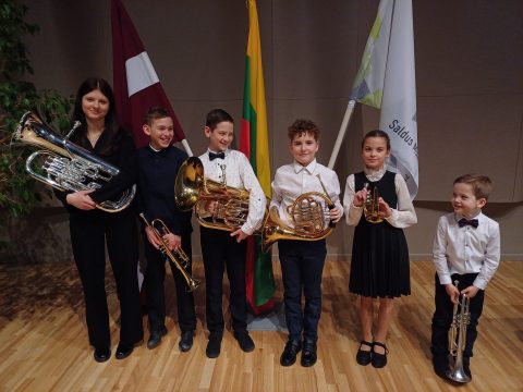 6 mūsu audzēkņi, turot rokās savus mūzikas instrumentus. Aizmugurē Latvijas, Lietuvas un Saldus Mūzikas skolas karogi.