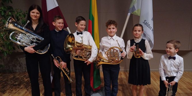 6 mūsu audzēkņi, turot rokās savus mūzikas instrumentus. Aizmugurē Latvijas, Lietuvas un Saldus Mūzikas skolas karogi.